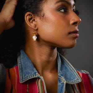 May include: A close-up of a person wearing a pair of skull earrings. The earrings feature a small white skull pendant with gold-colored accents. The person is wearing a denim vest over a colorful striped top.