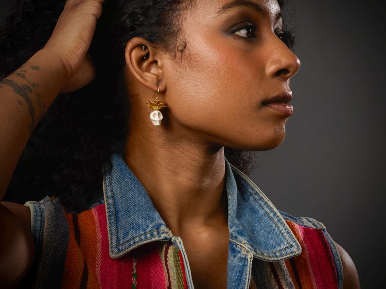 May include: A close-up of a person wearing a pair of skull earrings. The earrings feature a small white skull pendant with gold-colored accents. The person is wearing a denim vest over a colorful striped top.