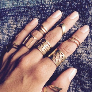 Op de afbeelding: Een hand met meerdere gouden ringen met geometrische ontwerpen. De ringen zijn op elke vinger gestapeld en zijn gemaakt van dun metaal.