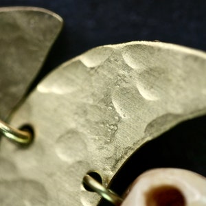 May include: Close-up of a gold crescent moon-shaped earring with a textured surface. The earring is attached to a white bone-like bead.