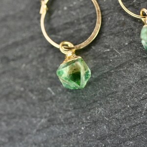 Puede incluir: Primer plano de dos pendientes de aro dorados, cada uno con un colgante de piedra preciosa verde facetada. Las piedras preciosas tienen una mezcla de tonos verdes claros y oscuros. Los pendientes est&aacute;n sobre un fondo oscuro y texturizado.