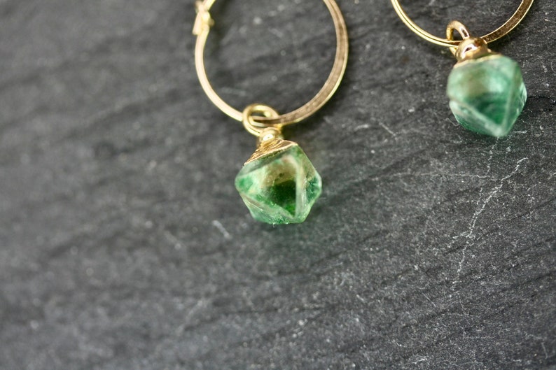 Puede incluir: Primer plano de dos pendientes de aro dorados, cada uno con un colgante de piedra preciosa verde facetada. Las piedras preciosas tienen una mezcla de tonos verdes claros y oscuros. Los pendientes est&aacute;n sobre un fondo oscuro y texturizado.