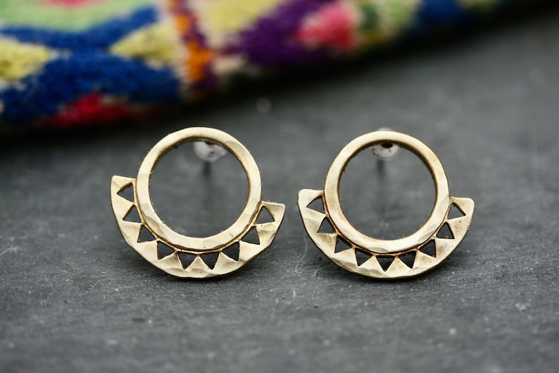 May include: A pair of gold-toned earrings with a hammered finish. Each earring features a circle with a half-circle of triangular cutouts around the bottom.
