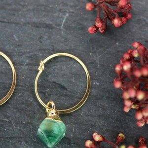 Puede incluir: Pendientes de aro dorados con un colgante de piedra verde facetada. Los pendientes se exhiben sobre una superficie gris oscuro, con una ramita de bayas rojas en el fondo. Joyas.