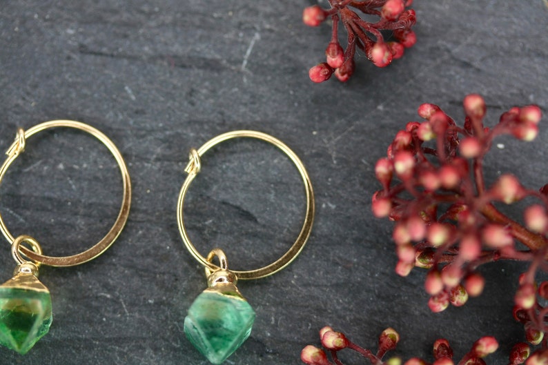 Puede incluir: Pendientes de aro dorados con un colgante de piedra verde facetada. Los pendientes se exhiben sobre una superficie gris oscuro, con una ramita de bayas rojas en el fondo. Joyas.