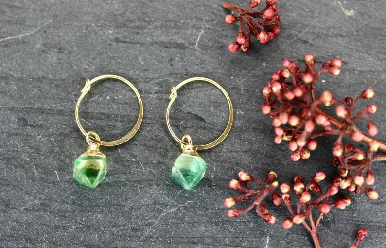 Puede incluir: Un par de pendientes de aro dorados con colgantes de piedras preciosas verdes facetadas. Los pendientes se exhiben sobre una superficie gris oscuro, con ramitas de bayas rojas en el fondo. La joyer&iacute;a es elegante y adecuada para el uso diario.