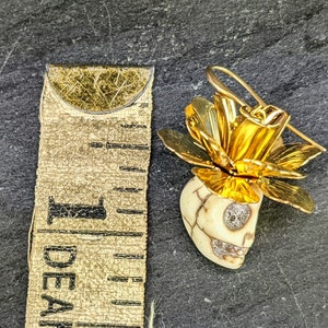 May include: A gold-colored floral earring with a skull-shaped pendant. The skull is off-white with a cracked pattern and sparkling accents in the eyes and mouth. The earring measures approximately 1 inch in length.