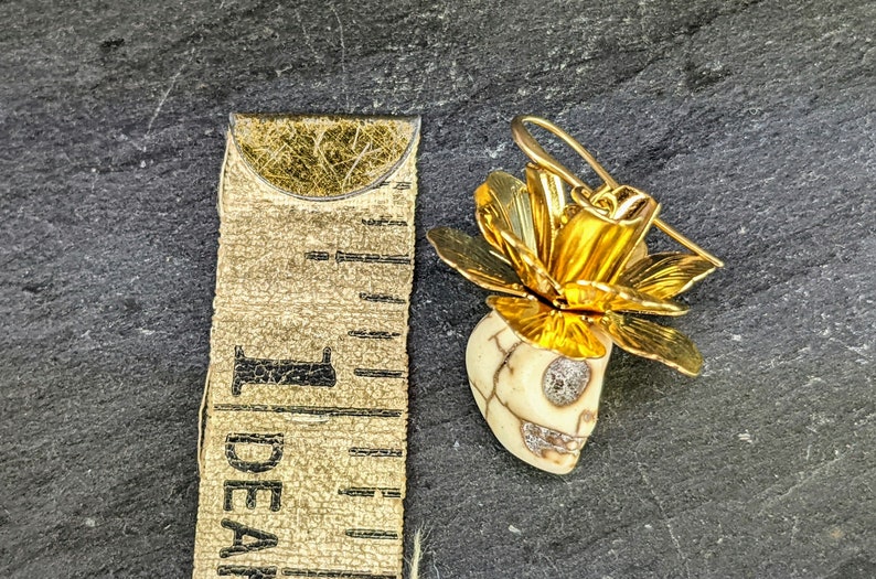 May include: A gold-colored floral earring with a skull-shaped pendant. The skull is off-white with a cracked pattern and sparkling accents in the eyes and mouth. The earring measures approximately 1 inch in length.