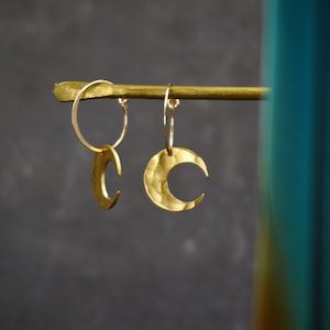 May include: Gold-colored crescent moon earrings. The earrings are hanging from a gold bar. The moons have a hammered texture. The background is a dark gray color.