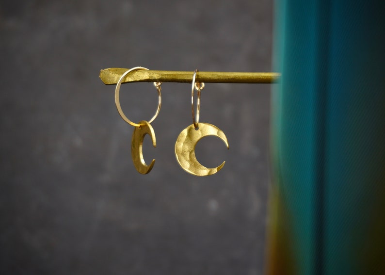 May include: Gold-colored crescent moon earrings. The earrings are hanging from a gold bar. The moons have a hammered texture. The background is a dark gray color.