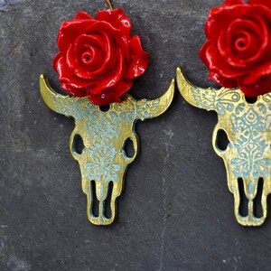 Puede incluir: Un par de pendientes de calavera de toro dorados con detalles de rosas rojas. Las calaveras de toro est&aacute;n detalladas con un patr&oacute;n floral azul y blanco.