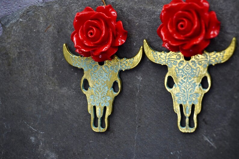 Puede incluir: Un par de pendientes de calavera de toro dorados con detalles de rosas rojas. Las calaveras de toro est&aacute;n detalladas con un patr&oacute;n floral azul y blanco.