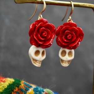 May include: A pair of dangle earrings with a white skull charm and a red rose charm. The earrings are made of metal and have a gold finish.