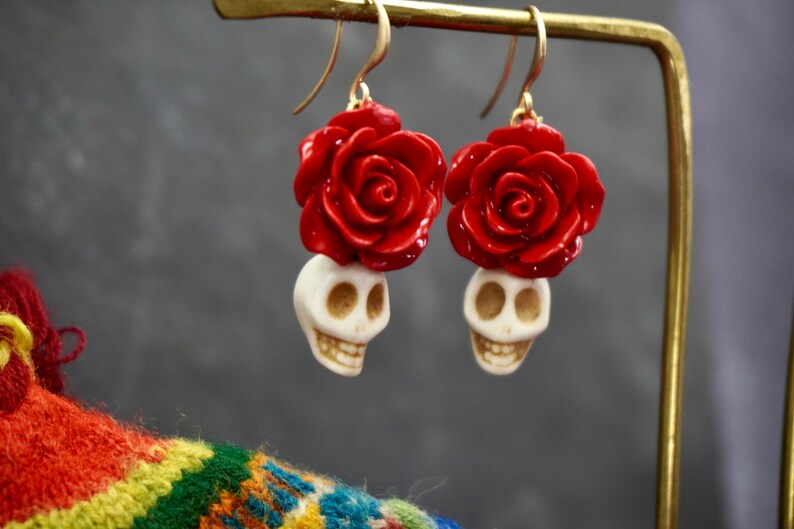 May include: A pair of dangle earrings with a white skull charm and a red rose charm. The earrings are made of metal and have a gold finish.