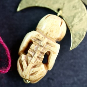 May include: A carved bone pendant with intricate details, hanging from a gold crescent moon charm. The pendant is a light beige color and has a smooth, polished finish.