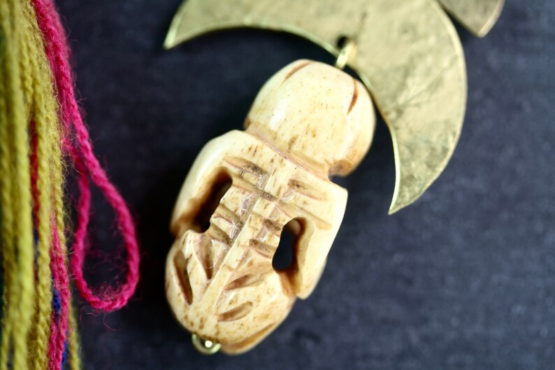 May include: A carved bone pendant with intricate details, hanging from a gold crescent moon charm. The pendant is a light beige color and has a smooth, polished finish.
