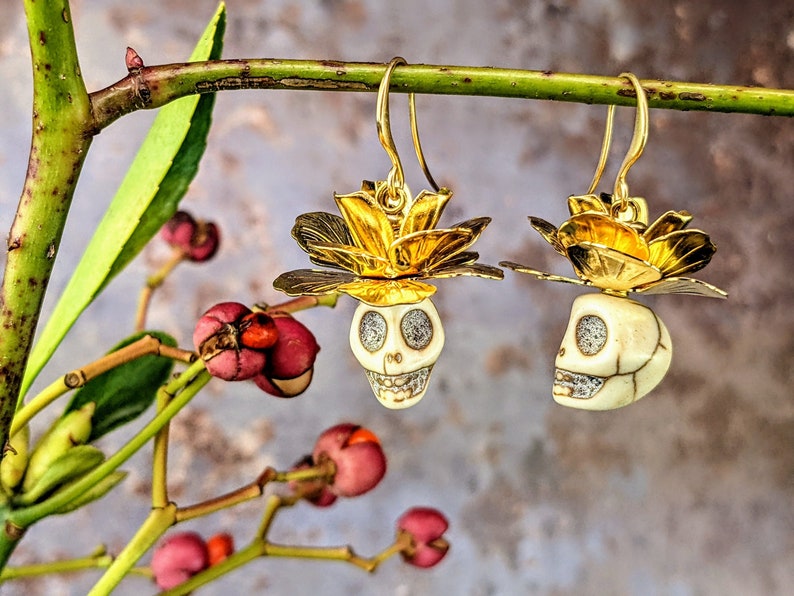 May include: A pair of gold-colored earrings featuring a skull design. The skulls are cream-colored with silver accents and are topped with a gold flower. The earrings hang from gold hooks and are displayed against a blurred background.