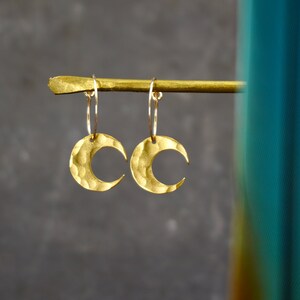 May include: A pair of gold-colored crescent moon earrings. Each earring features a hammered texture and hangs from a thin hoop. The earrings are displayed against a dark gray background.