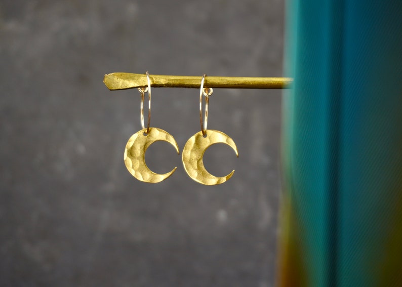 May include: A pair of gold-colored crescent moon earrings. Each earring features a hammered texture and hangs from a thin hoop. The earrings are displayed against a dark gray background.