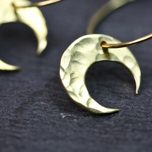 May include: Close-up of two gold-colored crescent moon earrings. The earrings have a hammered texture and are suspended from a thin, curved wire. The background is a dark, textured surface.