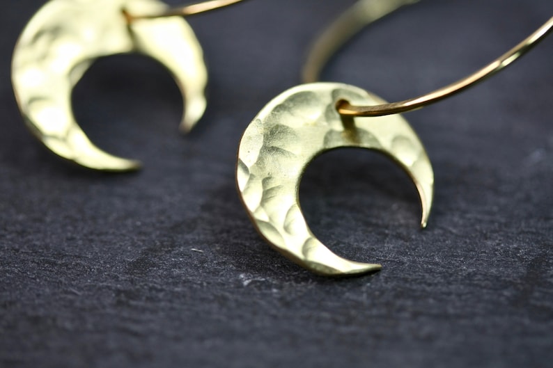 May include: Close-up of two gold-colored crescent moon earrings. The earrings have a hammered texture and are suspended from a thin, curved wire. The background is a dark, textured surface.