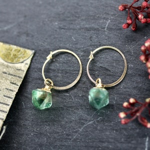 Puede incluir: Un par de pendientes de aro dorados con colgantes de piedras preciosas verdes. Los pendientes se muestran sobre una superficie gris oscuro, con una cinta m&eacute;trica y ramitas de bayas rojas en el fondo.