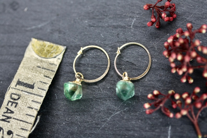 Puede incluir: Un par de pendientes de aro dorados con colgantes de piedras preciosas verdes. Los pendientes se muestran sobre una superficie gris oscuro, con una cinta m&eacute;trica y ramitas de bayas rojas en el fondo.