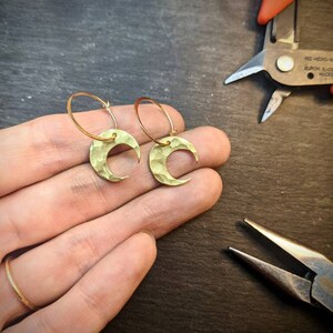 May include: A pair of gold-colored crescent moon earrings with textured surfaces. The earrings are on a gold hoop. The image also shows a hand with a gold ring and jewelry-making tools in the background.