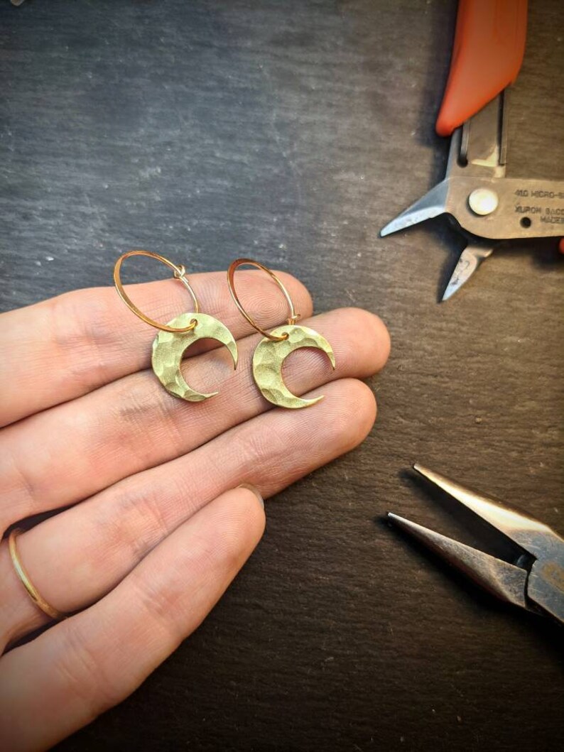 May include: A pair of gold-colored crescent moon earrings with textured surfaces. The earrings are on a gold hoop. The image also shows a hand with a gold ring and jewelry-making tools in the background.