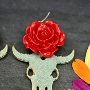 Puede incluir: Un par de pendientes con una rosa roja en la parte superior de un cr&aacute;neo de toro dorado con un dise&ntilde;o de patr&oacute;n azul y dorado.