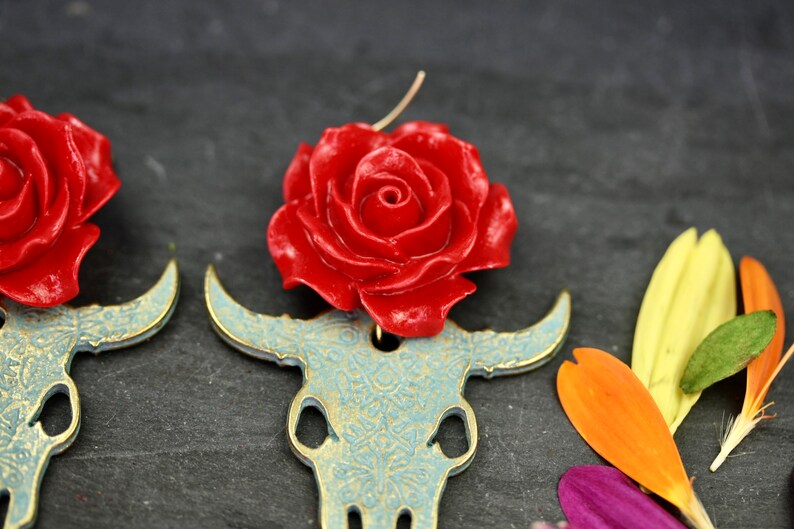 Puede incluir: Un par de pendientes con una rosa roja en la parte superior de un cr&aacute;neo de toro dorado con un dise&ntilde;o de patr&oacute;n azul y dorado.