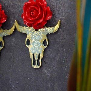 Puede incluir: Un par de pendientes de calavera de toro dorados con detalles de rosas rojas. Los pendientes est&aacute;n detallados con un patr&oacute;n floral blanco.