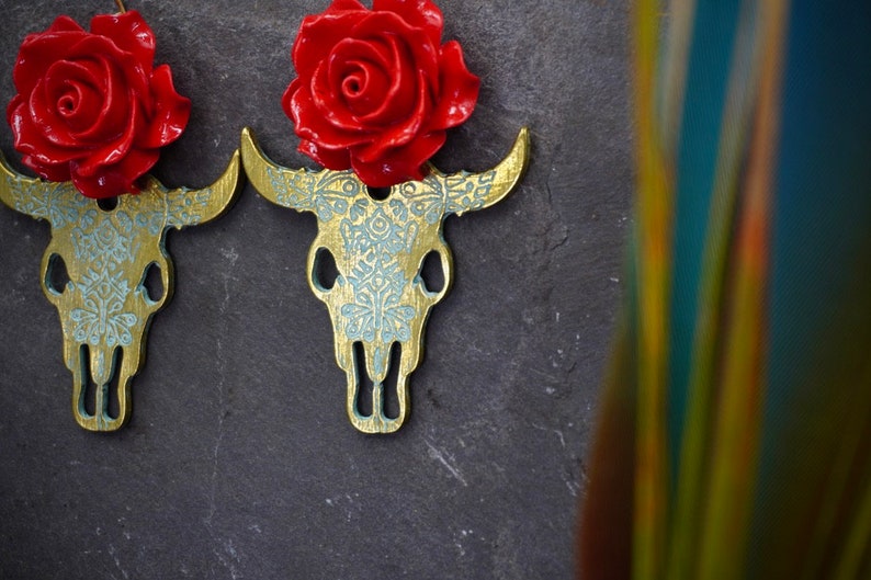 Puede incluir: Un par de pendientes de calavera de toro dorados con detalles de rosas rojas. Los pendientes est&aacute;n detallados con un patr&oacute;n floral blanco.