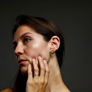 May include: A woman with long brown hair wearing gold earrings and a gold ring. She is looking to the side and touching her face with her hand.