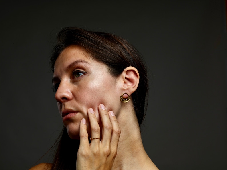 May include: A woman with long brown hair wearing gold earrings and a gold ring. She is looking to the side and touching her face with her hand.