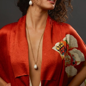 May include: A woman wearing a long gold necklace with a large pearl pendant. She is also wearing pearl earrings. The woman is wearing a red kimono with a floral design on the sleeve. The background is a dark gray.