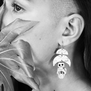 May include: A close-up of a person's face with a leaf covering part of their face. The person is wearing a pair of silver earrings with a skull charm.