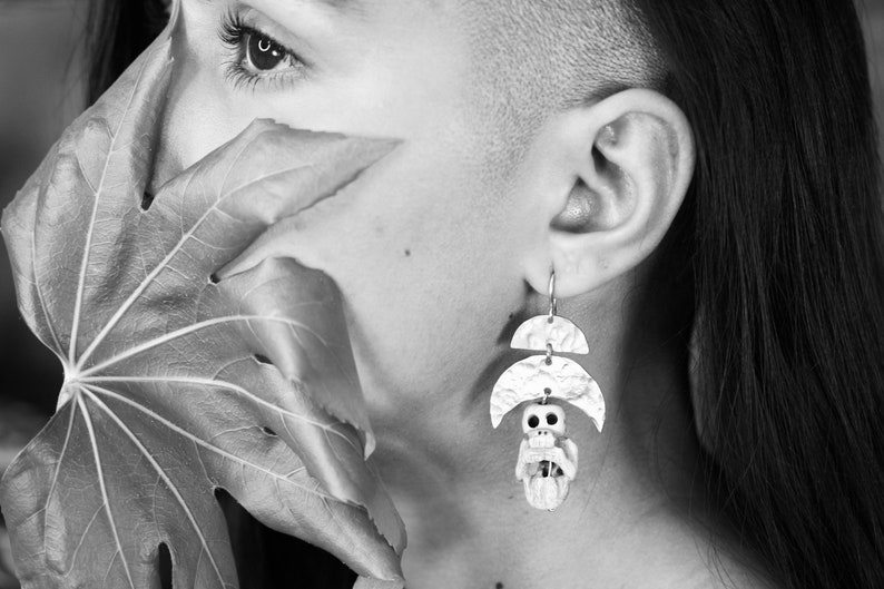 May include: A close-up of a person's face with a leaf covering part of their face. The person is wearing a pair of silver earrings with a skull charm.