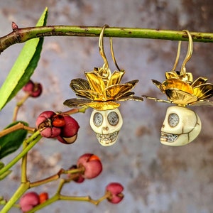 May include: Gold-tone earrings featuring skull pendants with flower accents. The skull pendants are off-white with detailed facial features and sparkling eyes. The earrings are suspended from gold-tone hooks, showcasing a unique and artistic design.
