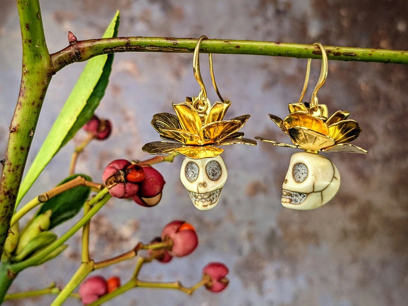 May include: Gold-tone earrings featuring skull pendants with flower accents. The skull pendants are off-white with detailed facial features and sparkling eyes. The earrings are suspended from gold-tone hooks, showcasing a unique and artistic design.