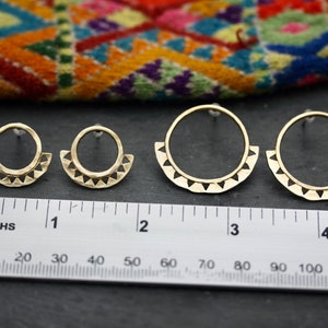 May include: Three pairs of gold-toned earrings with a geometric design. The earrings are in different sizes, with the largest pair measuring approximately 7.5 cm in diameter. The earrings are shown on a black surface with a ruler for scale.