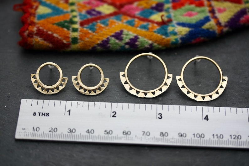 May include: Three pairs of gold-toned earrings with a geometric design. The earrings are in different sizes, with the largest pair measuring approximately 7.5 cm in diameter. The earrings are shown on a black surface with a ruler for scale.
