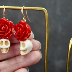 May include: A pair of dangle earrings featuring a white skull with a red rose on top. The earrings are made of polymer clay and have gold hooks.