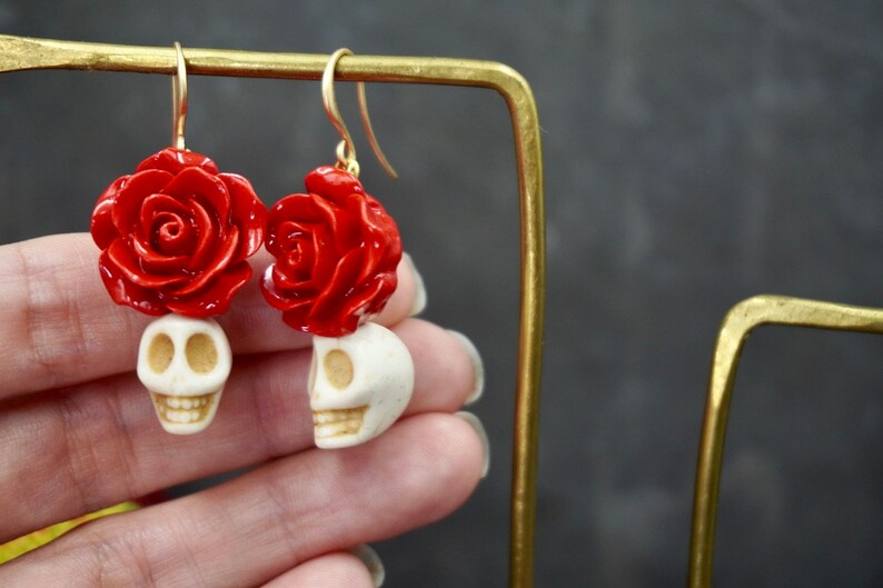 May include: A pair of dangle earrings featuring a white skull with a red rose on top. The earrings are made of polymer clay and have gold hooks.