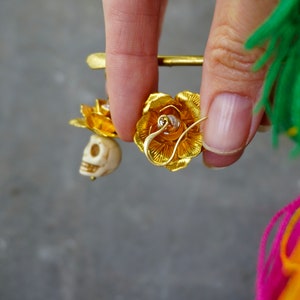 May include: Gold-colored earrings featuring a rose design with a small skull charm. The earrings are held up by a gold pin. The rose petals are detailed, and the skull is off-white. The background is a blurred gray.