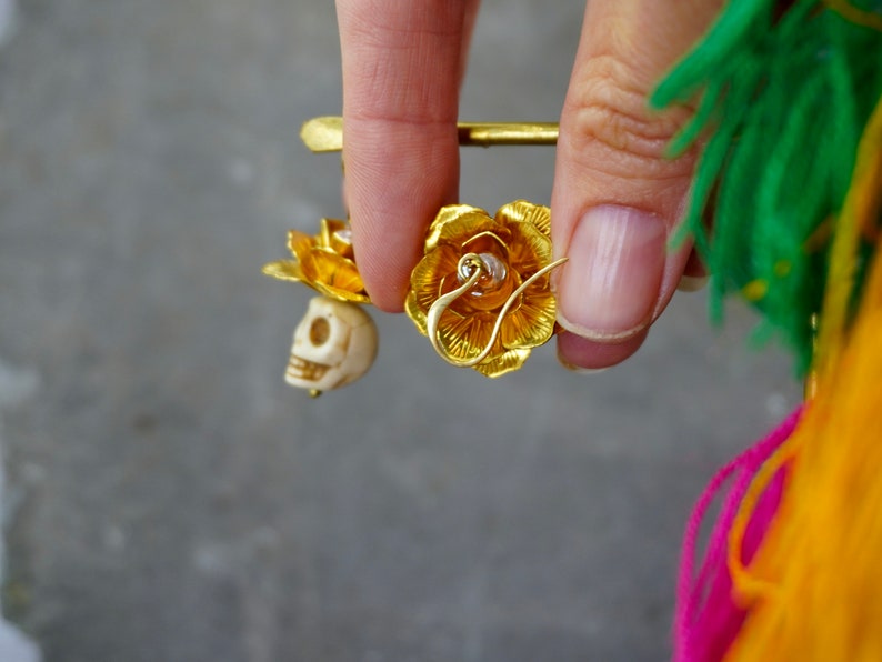 May include: Gold-colored earrings featuring a rose design with a small skull charm. The earrings are held up by a gold pin. The rose petals are detailed, and the skull is off-white. The background is a blurred gray.