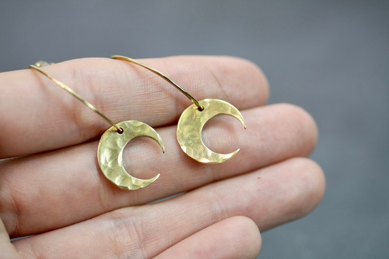 May include: A pair of gold-colored crescent moon earrings. The earrings have a hammered texture and are suspended from delicate, curved wires. The design is simple and elegant, suitable for everyday wear or special occasions.