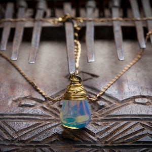 May include: A gold chain necklace with a teardrop-shaped pendant. The pendant is made of a blue and white iridescent stone wrapped in gold wire. The necklace is lying on a wooden surface with a carved pattern.