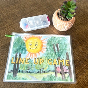 May include: A clear plastic storage container with a red lid and a colorful assortment of small objects inside. The container is sitting on a wooden table next to a potted succulent plant with green leaves and a terracotta pot with a decorative pattern. A clear plastic sheet with a watercolor illustration of a smiling sun, trees, and a red barn with the text "LINE UP GAME" is on the table.