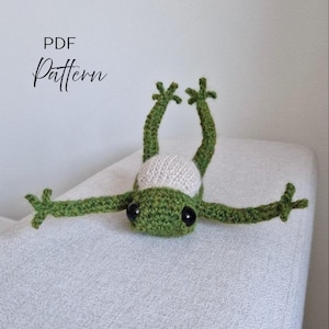 May include: A green and cream-colored crocheted frog toy with large black plastic eyes, positioned with its legs extended upward and arms stretched out to the sides. The text PDF Pattern is visible in the upper left corner.